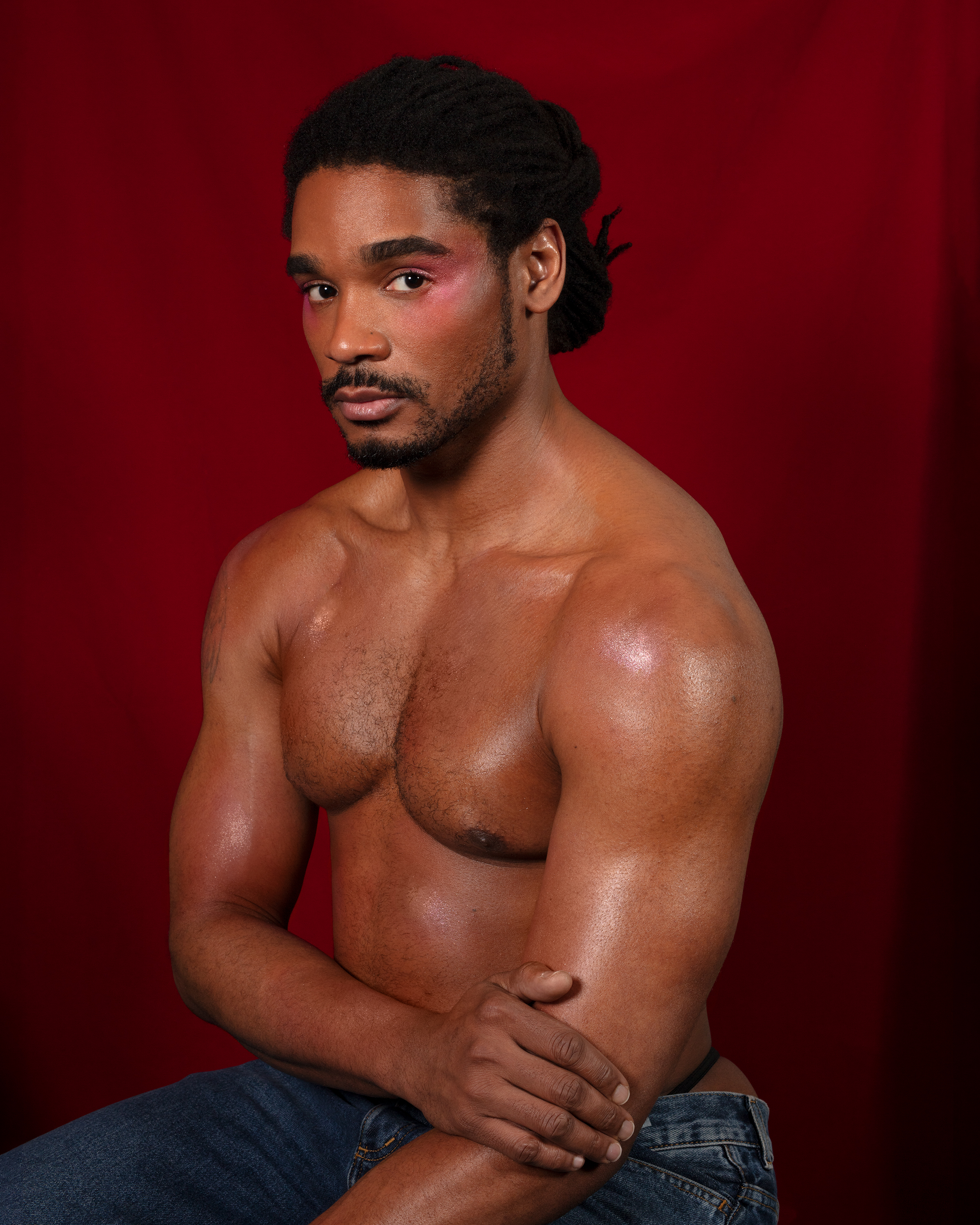 Waist up portrait of Khalyle shirtless with glowing skin looking at the camera against a red backdrop